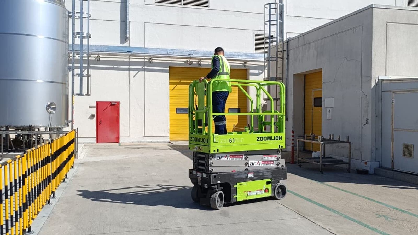 Fabrika bahçesinde dar geçitte konumlanmış kompakt akülü makaslı platform ve reflektif yelekli operatör